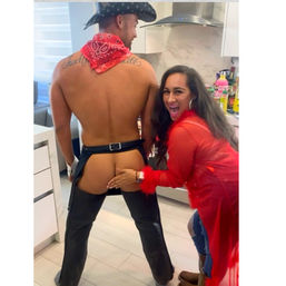 Laughing woman playfully touching a shirtless man in a cowboy hat and chaps with exposed butt during a lively kitchen party