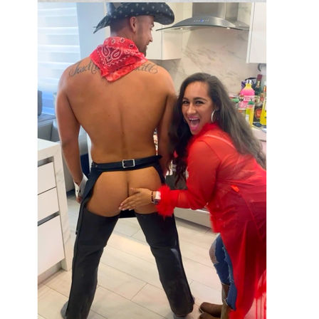Laughing woman playfully touching a shirtless man in a cowboy hat and chaps with exposed butt during a lively kitchen party