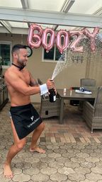 Shirtless man in black apron and cuff popping a champagne bottle, spraying bubbly across a backyard patio with rose-gold letter balloons and wicker dining set.