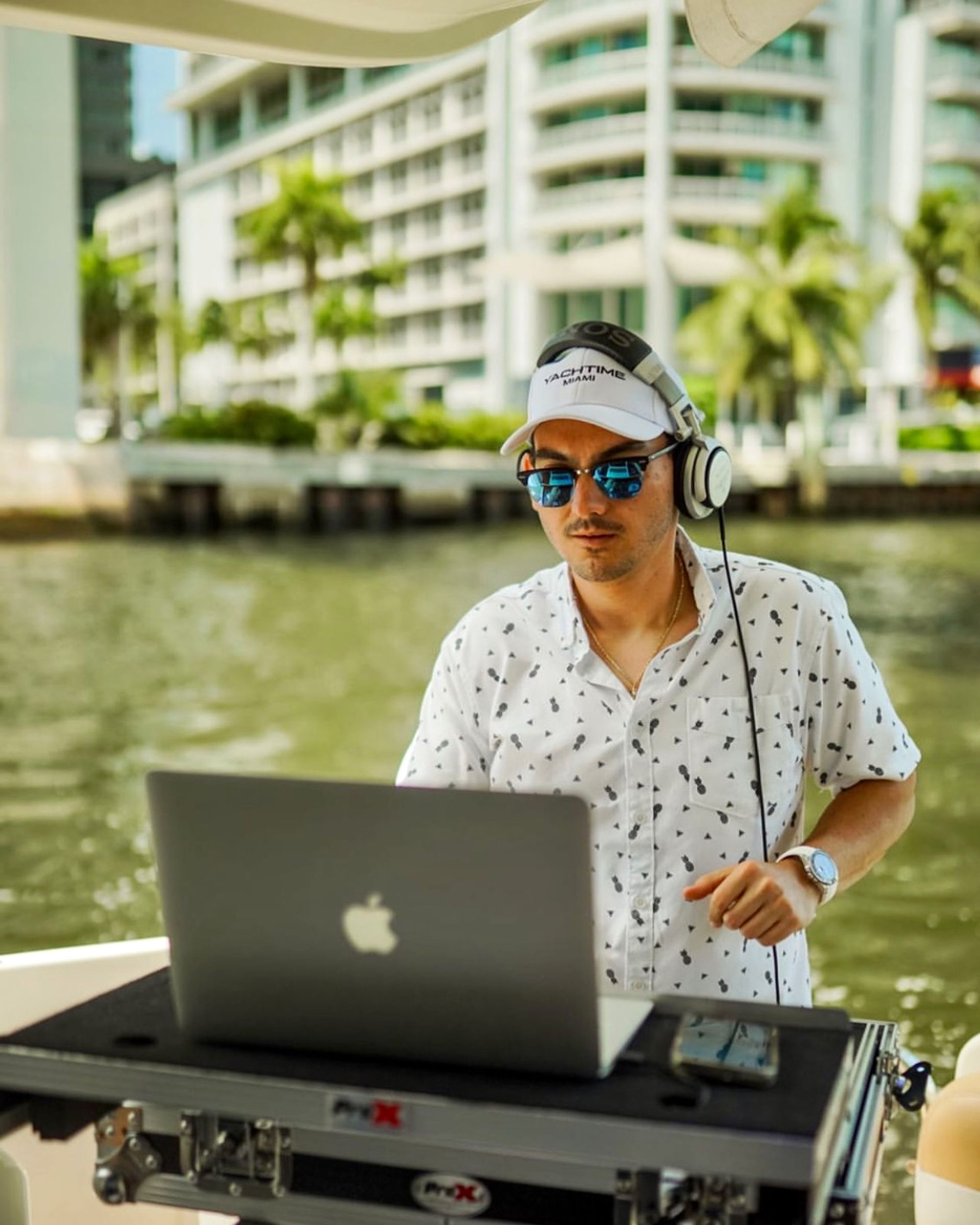 DJ in sunglasses and headphones mixing on a laptop aboard a yacht on the Miami waterfront with palm trees and beachfront condos in the background.