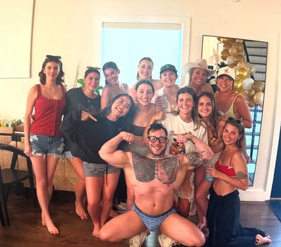 Group of friends posing in a sunlit living room at a casual summer house party — women in shorts, swimsuits and hats and a tattooed man flexing in patterned briefs in front, with gold-and-white balloons reflected in a mirror.