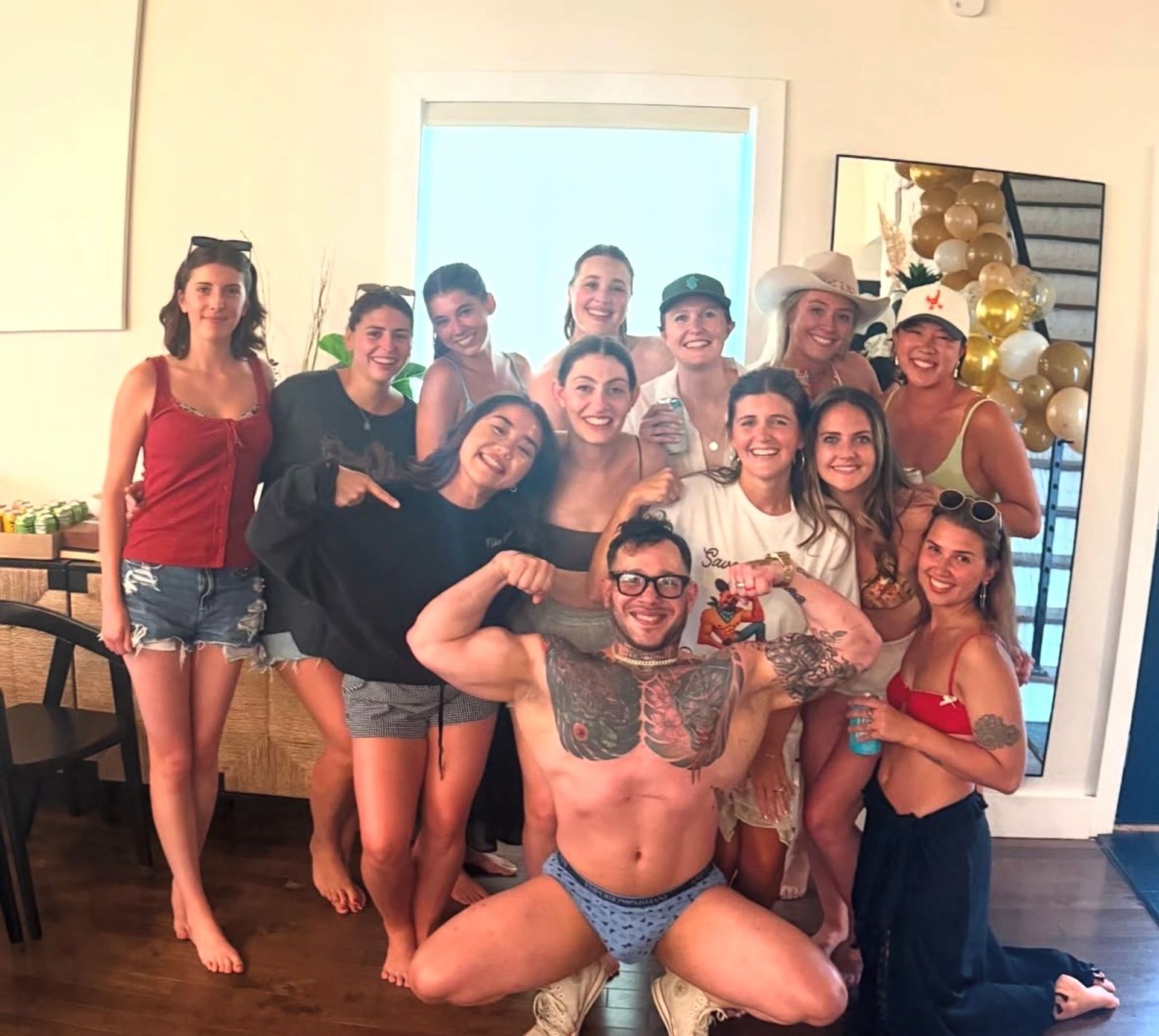 Group of friends posing in a sunlit living room at a casual summer house party — women in shorts, swimsuits and hats and a tattooed man flexing in patterned briefs in front, with gold-and-white balloons reflected in a mirror.