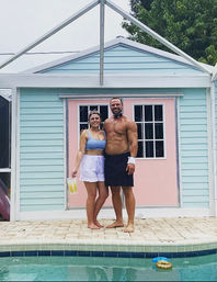 Smiling adult couple in swimwear posing poolside in front of a pastel blue-and-pink pool cabana by a backyard swimming pool.