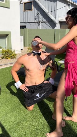 Sunny backyard poolside scene — shirtless man kneeling on artificial turf wearing a bow tie and cuffs while a woman in a pink cover-up pours water into his mouth.