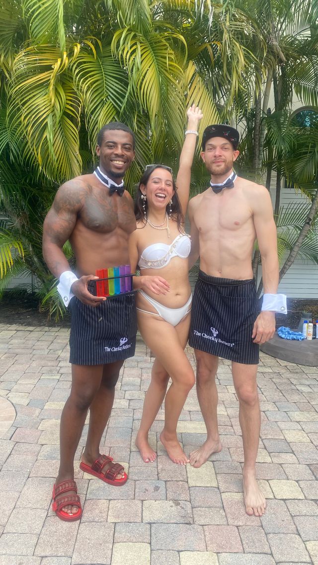 Tropical poolside party on a paver patio: laughing woman in a white bikini and pearls between two shirtless bow-tied servers in aprons, one holding a rainbow shot flight, palm trees behind.