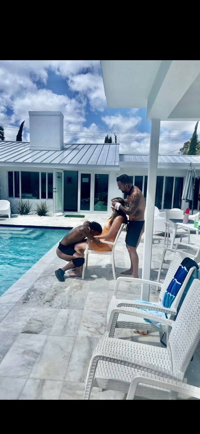 Sunny poolside scene at a modern white house: two shirtless men and a woman in an orange dress on a marble patio beside a turquoise pool, one man kneeling at her feet and the other standing behind gently touching her head, white wicker chairs and a blue-striped towel nearby under a partly cloudy sky.