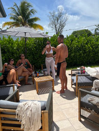 Sunny tropical patio with a palm tree and striped umbrella, friends in swimwear relaxing on wooden lounge chairs and sipping drinks while socializing outdoors.