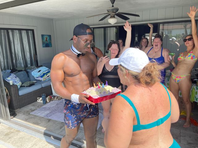 Muscular male entertainer in a bow tie hands a birthday cake to a woman in a turquoise swimsuit while a group of friends in swimsuits cheer on a sunny poolside patio.