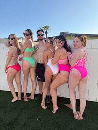 Group of six friends in colorful swimsuits posing barefoot against a white backyard wall under a clear blue sky with a palm tree, playful sunny pool-party vibe.