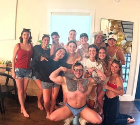 Group photo at an indoor summer party: a shirtless tattooed man flexes in patterned briefs while a dozen smiling women in swimsuits and casual summer outfits pose behind near a mirror and gold balloons.