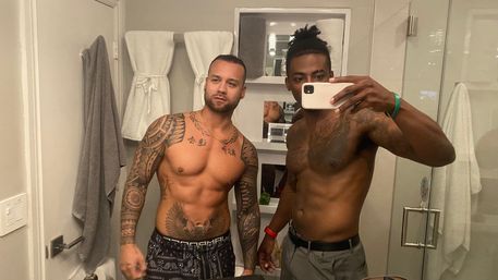 Bathroom mirror selfie of two shirtless, tattooed men posing with a smartphone in a modern white bathroom with towels, shelving, and a glass shower.