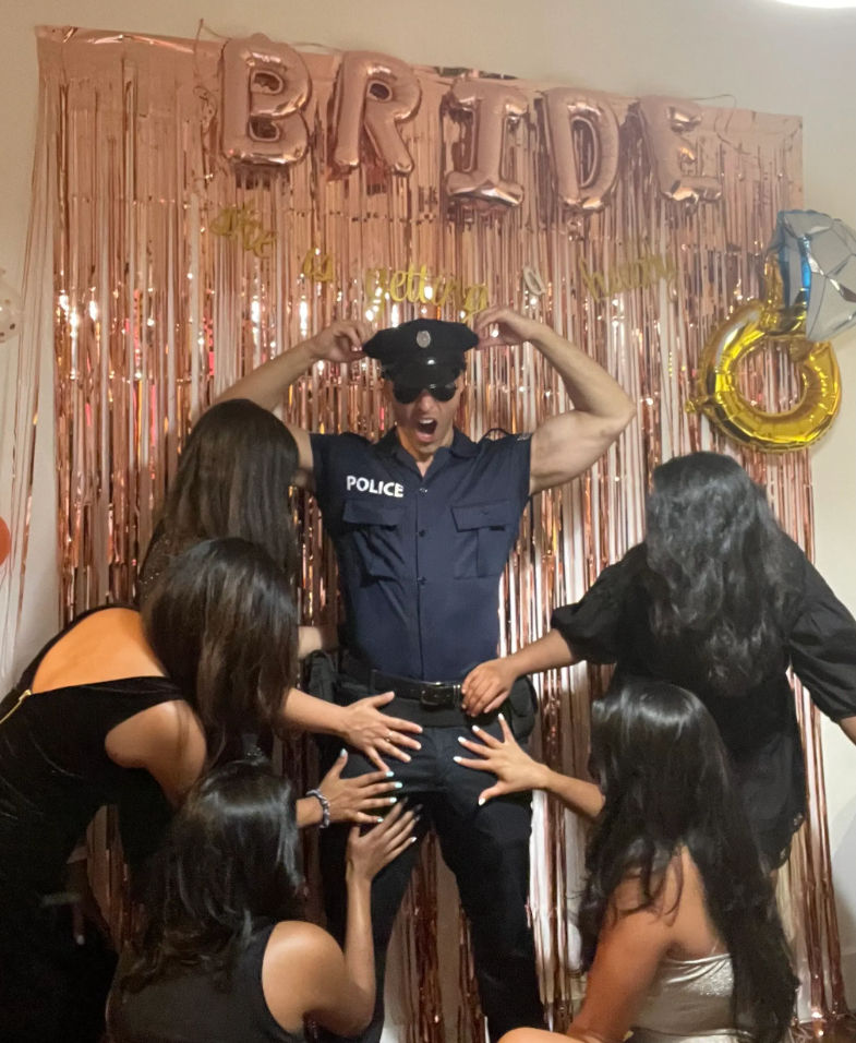 Bachelorette party scene with a man in a police costume striking a playful pose in front of a rose-gold fringe backdrop and 'BRIDE' balloons while several women in party dresses reach toward him