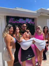 Sunlit backyard pool party: shirtless man in sunglasses lifts a woman wrapped in a white towel while friends in colorful swimsuits cheer and one sips champagne in front of a neon pink sign, green hedge and pink inflatable chair under a clear blue sky.