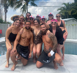 Poolside group photo at a tropical backyard party — smiling friends in swimsuits, sun hats and heart-shaped sunglasses clustered behind two shirtless men kneeling in striped aprons, palm trees and cloudy sky in the background.