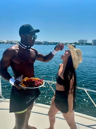 Playful scene on a sunny boat deck: shirtless man in cap, sunglasses and waiter-style cuffs offers grapes from a fruit platter to a woman in a straw hat and black bikini, with calm blue water and coastal buildings in the background.