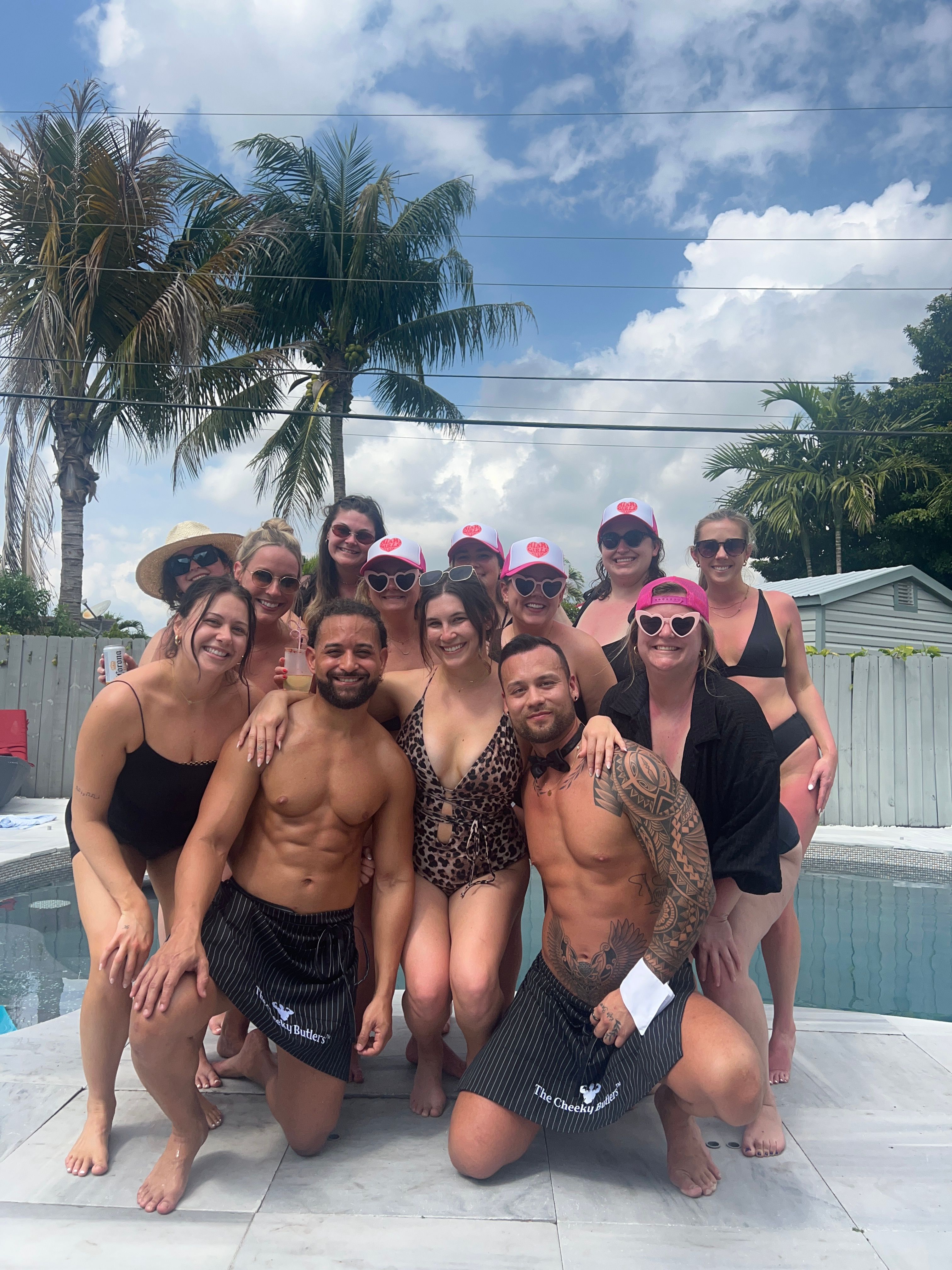 Smiling group of friends posing at a tropical backyard pool with two shirtless men kneeling in front, women in swimsuits and pink caps, palm trees and blue sky overhead.