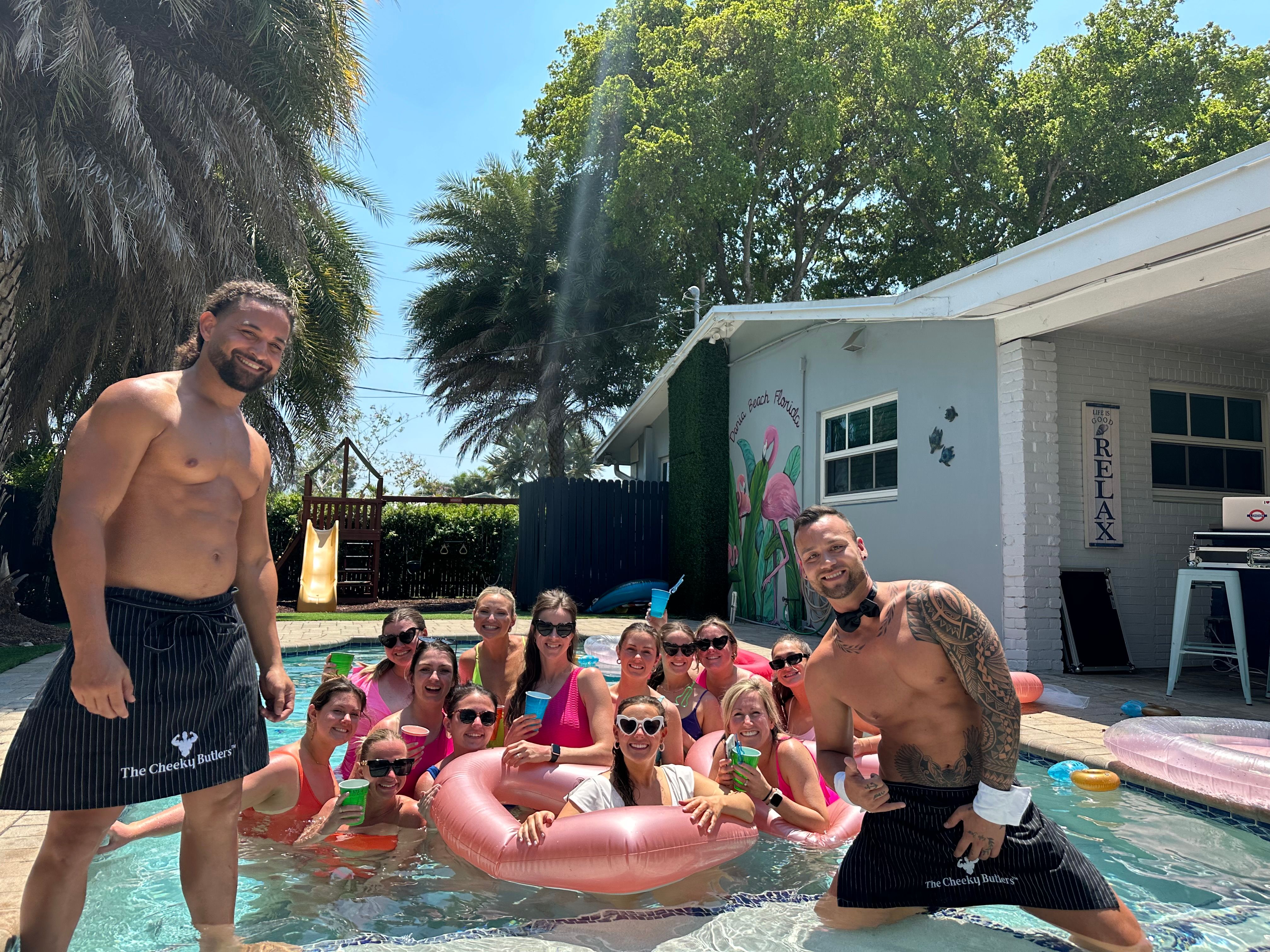 Sunny Florida backyard pool party: group of women relaxing in pink inflatable rings with colorful drinks, two shirtless male hosts posing at the pool edge, palm trees and a flamingo mural on the house wall.
