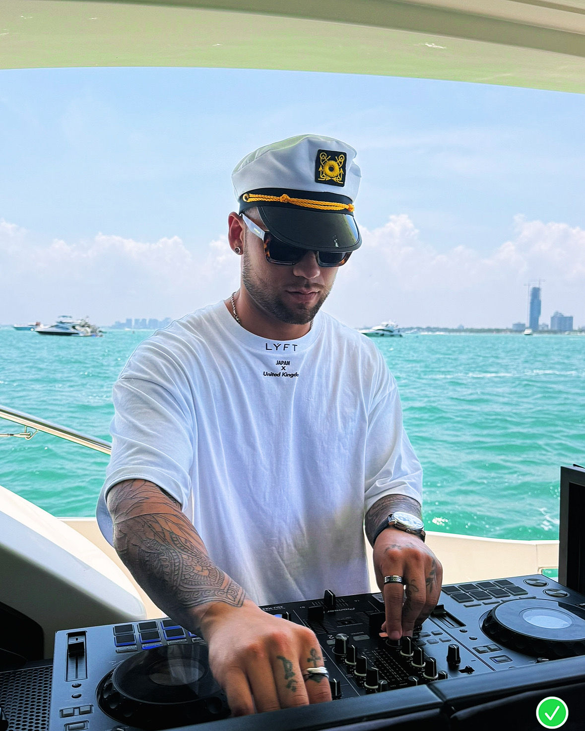 DJ wearing a captain's hat and sunglasses mixing on a yacht deck, tattooed arms and white tee against turquoise ocean and distant coastal skyline under a blue sky.