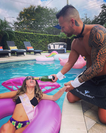 Backyard pool bachelorette: woman on pink inflatable float wearing sash drinks champagne poured by a shirtless bow-tie attendant with tattooed arm, pool loungers and inflatables in the background.
