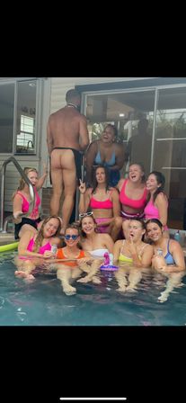 Backyard pool party: a group of people in colorful swimsuits laughing by a patio sliding door while a shirtless man in an apron stands with his bare backside visible.