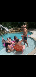 Backyard pool party with friends lounging in the shallow end on a pink inflatable and orange pool chair, one man standing in swim trunks taking a shot, others sipping drinks near a white water slide and rock waterfall surrounded by tropical greenery.