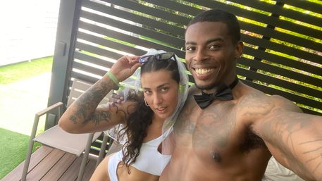 Smiling shirtless man in a black bow tie and tattoos takes a selfie with a woman in a white swimsuit and sheer veil, lounging in a shaded poolside cabana with chairs and green lawn visible.