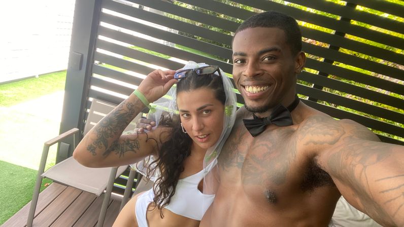 Smiling shirtless man in a black bow tie and tattoos takes a selfie with a woman in a white swimsuit and sheer veil, lounging in a shaded poolside cabana with chairs and green lawn visible.