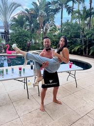 Tropical backyard pool party: man in bow tie and apron lifts laughing woman in heels near inflatable flamingo, folding tables with drinks and palm trees