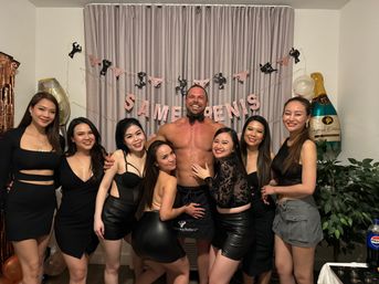 Bachelorette party in a living room: seven women in black party outfits smiling and posing around a muscular shirtless male entertainer wearing a bow tie and apron, pink garland and champagne balloons decorating the background.