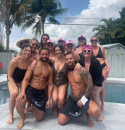 Smiling group at a tropical backyard pool: women in swimsuits, some with matching pink hats and heart-shaped sunglasses, posing with two shirtless men kneeling in front under palm trees.