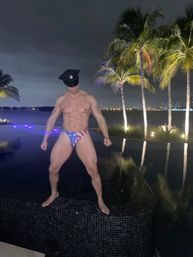 Shirtless muscular man in an American-flag swim brief and police cap striking a pose on the edge of a lit infinity pool at night, palm trees and distant waterfront city lights in the background.
