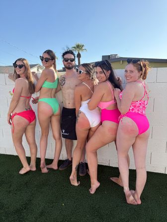 Group of six friends in colorful bikinis and swimwear posing barefoot against a white backyard wall under a clear blue sky with a palm tree, giving a fun sunny pool-party vibe.
