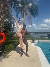 Two people in swimwear at a backyard lakeside pool — a man lifts a woman in heart-shaped sunglasses beside palm trees, string lights and a dock under a sunny blue sky.