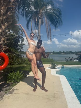 Two people in swimwear at a backyard lakeside pool — a man lifts a woman in heart-shaped sunglasses beside palm trees, string lights and a dock under a sunny blue sky.