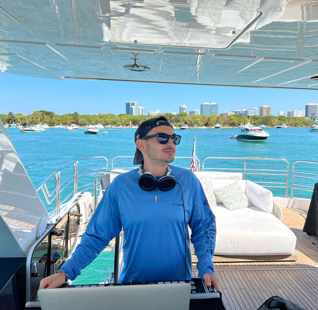 DJ with headphones and sunglasses on a luxury yacht deck, turquoise bay dotted with boats and a coastal city skyline under a clear blue sky — sunny waterfront party vibe.