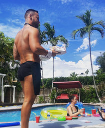 Shirtless pool server in black shorts carrying trays at a sunny tropical backyard pool party with palm trees, colorful inflatables and guests holding red cups.