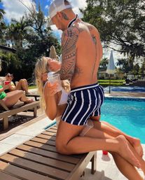 Playful poolside moment: tattooed man in a white cap and navy striped swim trunks kneels on a lounger while a woman in a white bikini laughs holding a clear cup, tropical palm trees and sunbathers in the sunny backyard pool background.