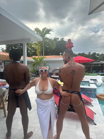 Playful poolside scene: smiling woman in white bikini and heart-shaped sunglasses posing between two men in cheeky swimwear by a tropical backyard pool with palm trees, red umbrellas and a cloudy sky.