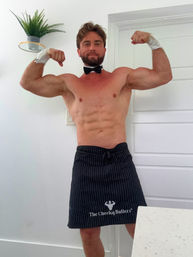Shirtless muscular man in a bow tie and black pinstripe apron playfully flexing biceps in a bright home kitchen.