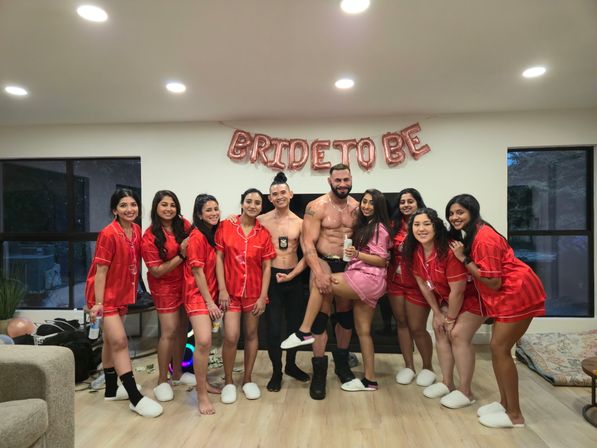 Bachelorette party group photo in a living room with rose-gold “BRIDE TO BE” balloons overhead; women in matching red satin pajamas and one in pink pose barefoot or in slippers with two shirtless male entertainers, hardwood floor and large windows in the background.
