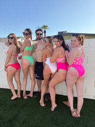 Six friends in colorful swimsuits and sunglasses posing against a white cinderblock wall in a sunny suburban backyard with a palm tree and blue sky, playful summer vibe.