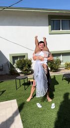 Playful scene of a man lifting a veiled woman upside down on green artificial turf in a sunny residential backyard, white stucco house with green-trim windows in the background.