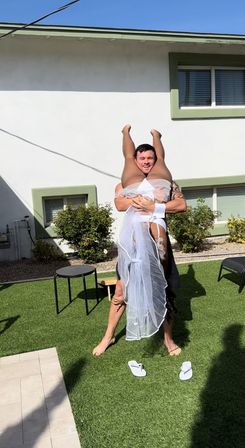 Playful scene of a man lifting a veiled woman upside down on green artificial turf in a sunny residential backyard, white stucco house with green-trim windows in the background.