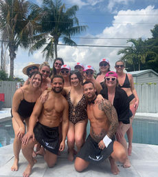 Smiling group of people in swimwear posing at a sunny backyard pool party with palm trees and blue sky in the background, some wearing pink caps and heart-shaped sunglasses.