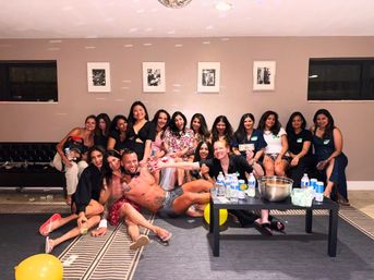 Smiling group of women at an indoor bachelorette-style party posing with a shirtless male entertainer in front of a couch, disco ball overhead, framed wall art, balloons on the floor and a table with water bottles and a punch bowl.