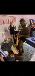 Bachelorette-style living room party: shirtless man in black briefs playfully entertaining a kneeling guest while friends cheer with drinks amid pink-and-gold balloons and tassel garland decorations.