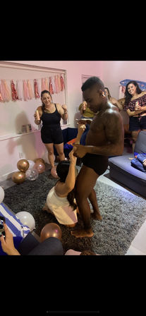 House party in a living room with pink tassel garland and rose-gold balloons, a shirtless man in black briefs dancing while a guest kneels and friends cheer with drinks