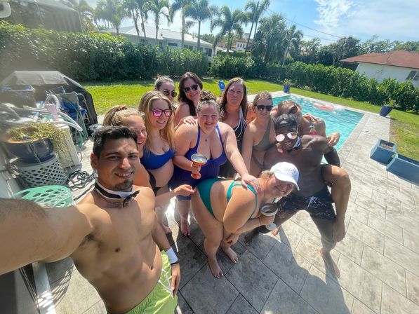 Sunlit backyard pool selfie of about a dozen adults in colorful swimsuits smiling and holding drinks at a palm‑lined suburban pool party.