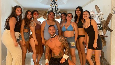Smiling group of women in swimsuits and summer dresses posing in a living room with a shirtless male entertainer kneeling in front wearing a bow tie and cuffs, chandelier and plants in the background — bachelorette party vibe
