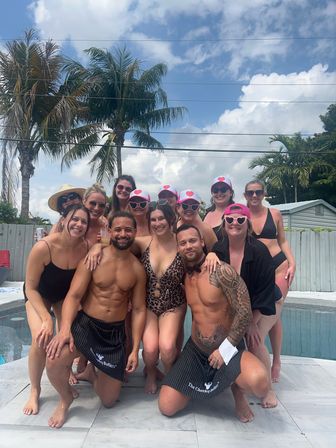 Fun tropical poolside group photo: adults in swimwear posing by a backyard pool with palm trees, blue sky, heart sunglasses and matching pink hats.